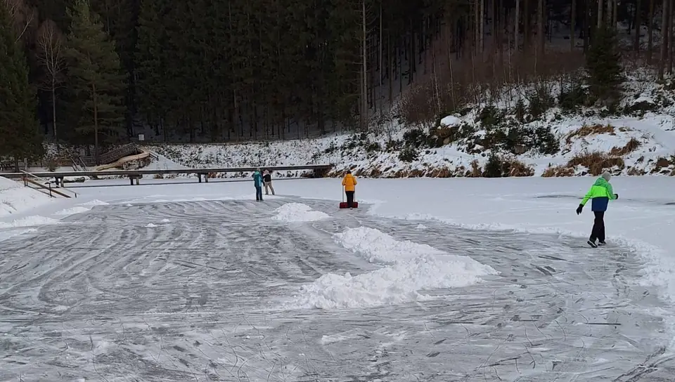 Geräumte Eisfläche auf dem zugefrorenen Waldbadteich mit Eisläufern, dahinter Holzsteg und Rutsche