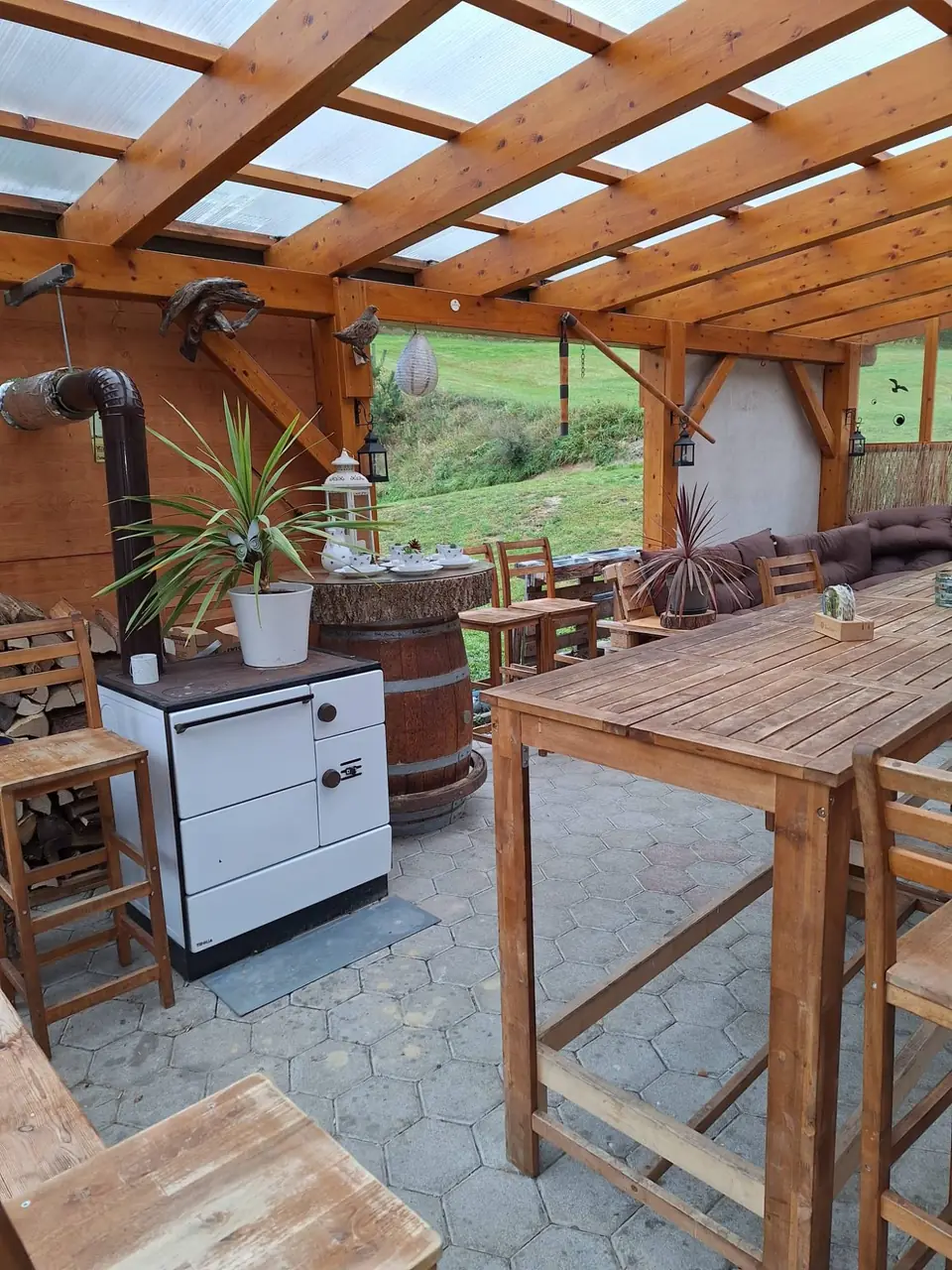 Gemütliche Ecke der überdachten Lounge mit Holzofen, Fass-Stehtisch und Brennholz