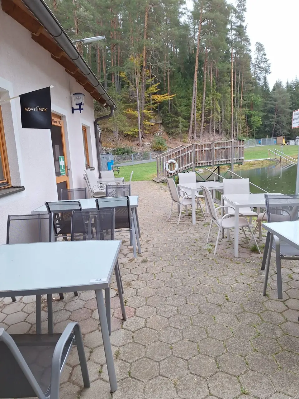 Terrasse mit Tischen und Stühlen direkt am Waldbadteich, daneben der Sprungturm